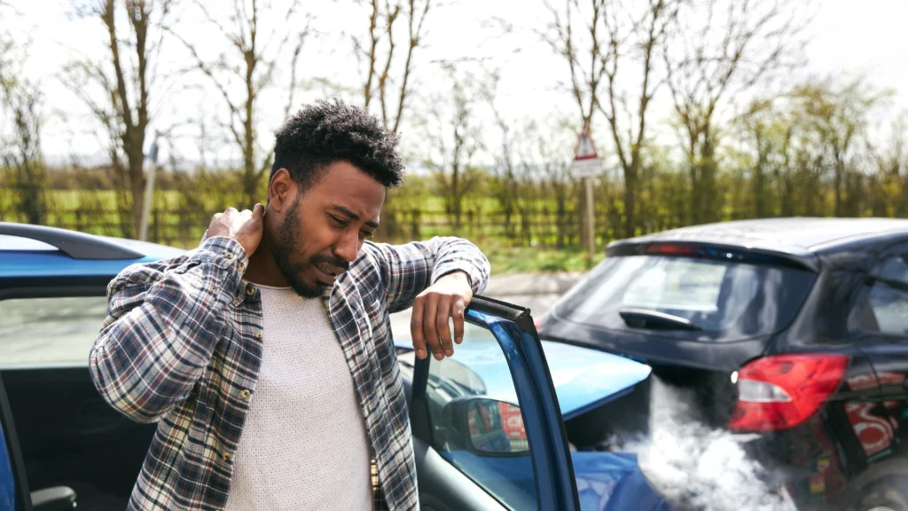 A person standing next to a car with smoke, dealing with the aftermath of an accident, potentially involving property damage liability