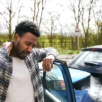A person standing next to a car with smoke, dealing with the aftermath of an accident, potentially involving property damage liability